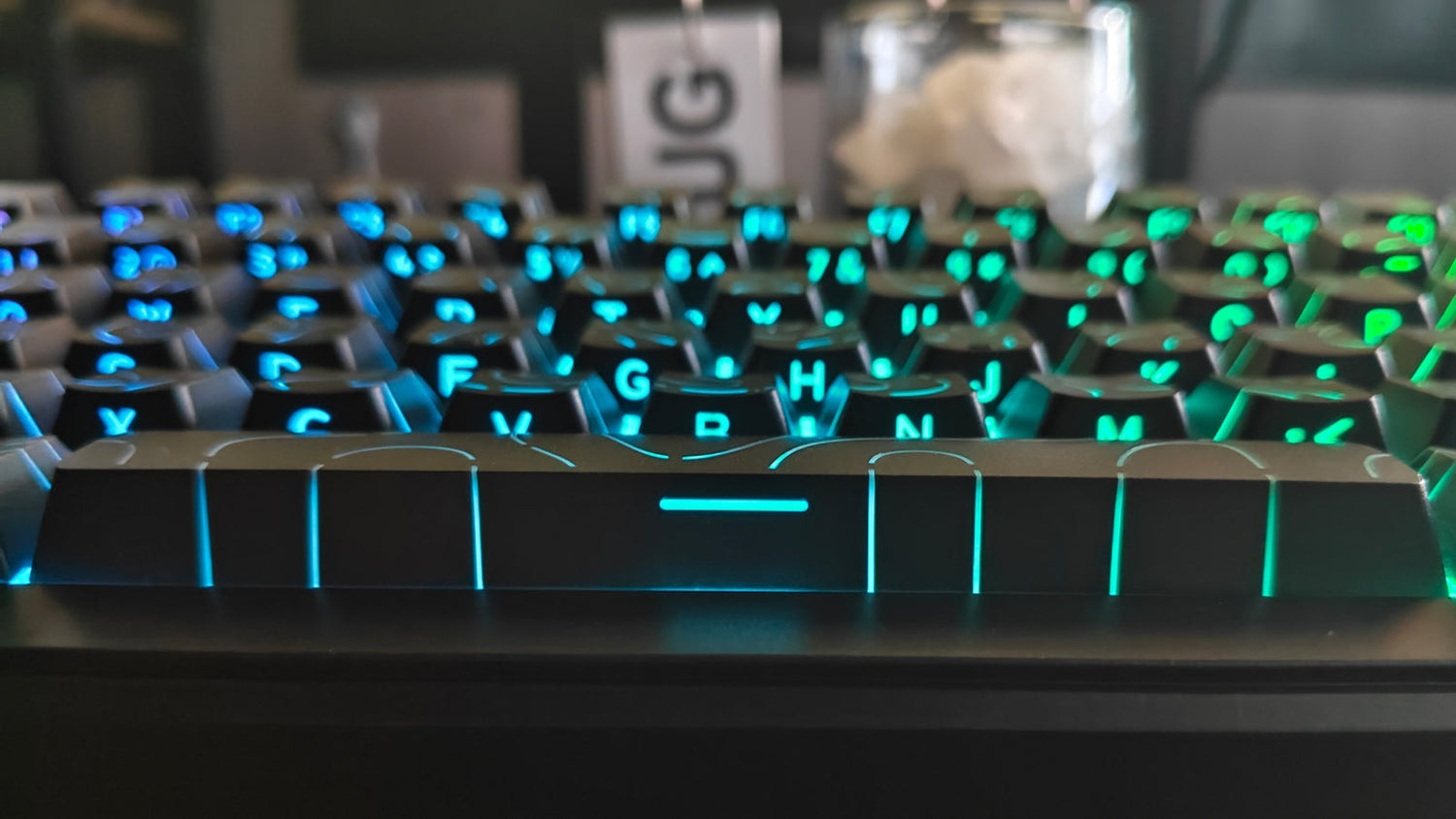 A close-up of a black gaming keyboard with blue and green backlighting, focusing on the spacebar