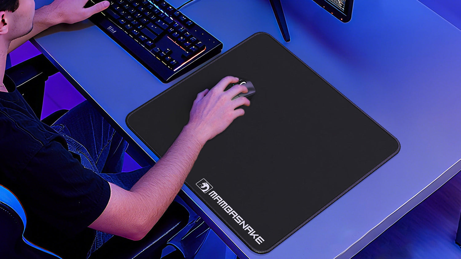 A person's hand using a computer mouse on a large black Mambasnake mousepad, placed on a blue desk next to a keyboard