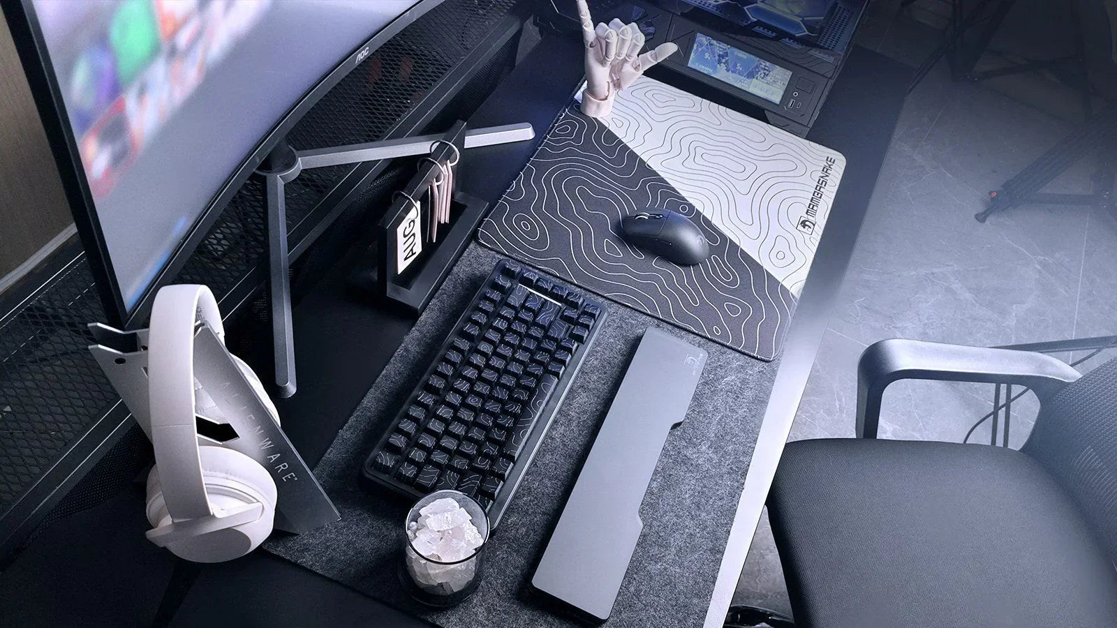 A modern, monochromatic gaming desk setup with a curved monitor, mechanical keyboard, white headphones, and a topographic mousepad