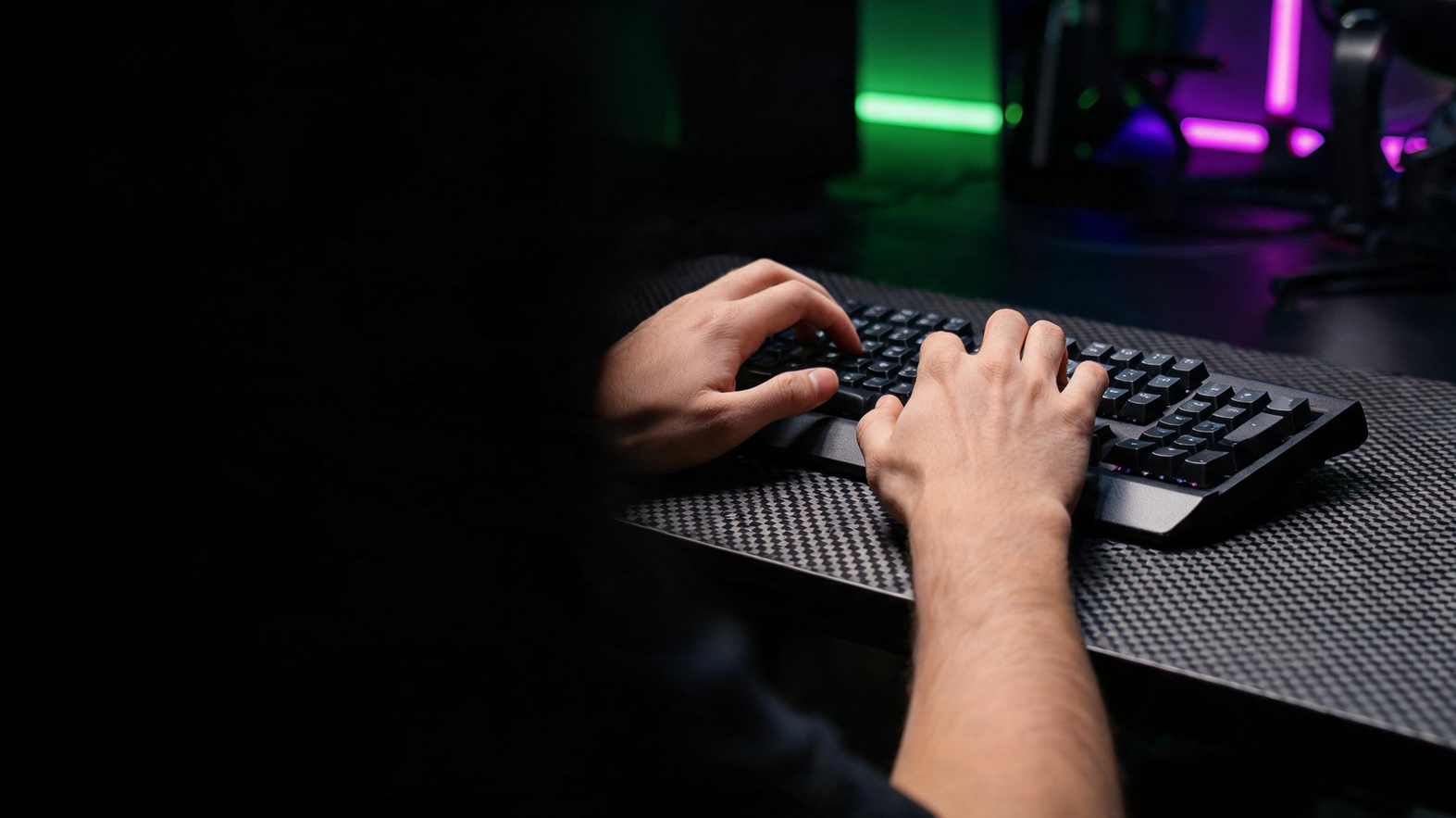 Gamer’s hands rest over a keyboard on a carbon-fiber desk in a neon-lit gaming room, with one side of the frame left dark.