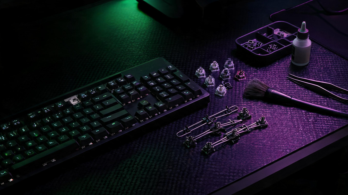 Mechanical keyboard with several switches and wire stabilizers arranged beside a fine brush and lubricant on a dark gaming desk