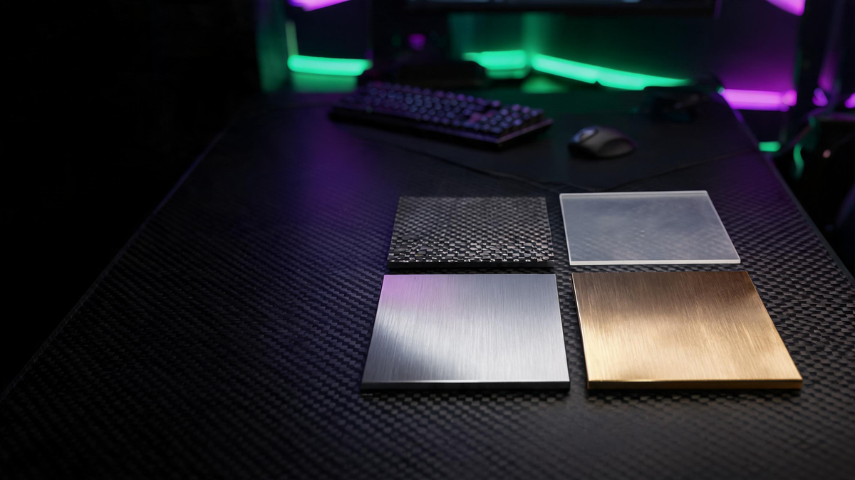 Four switch plate material samples—FR4, translucent polycarbonate, aluminum, and brass—arranged on a carbon-fiber gaming desk under green and purple lighting