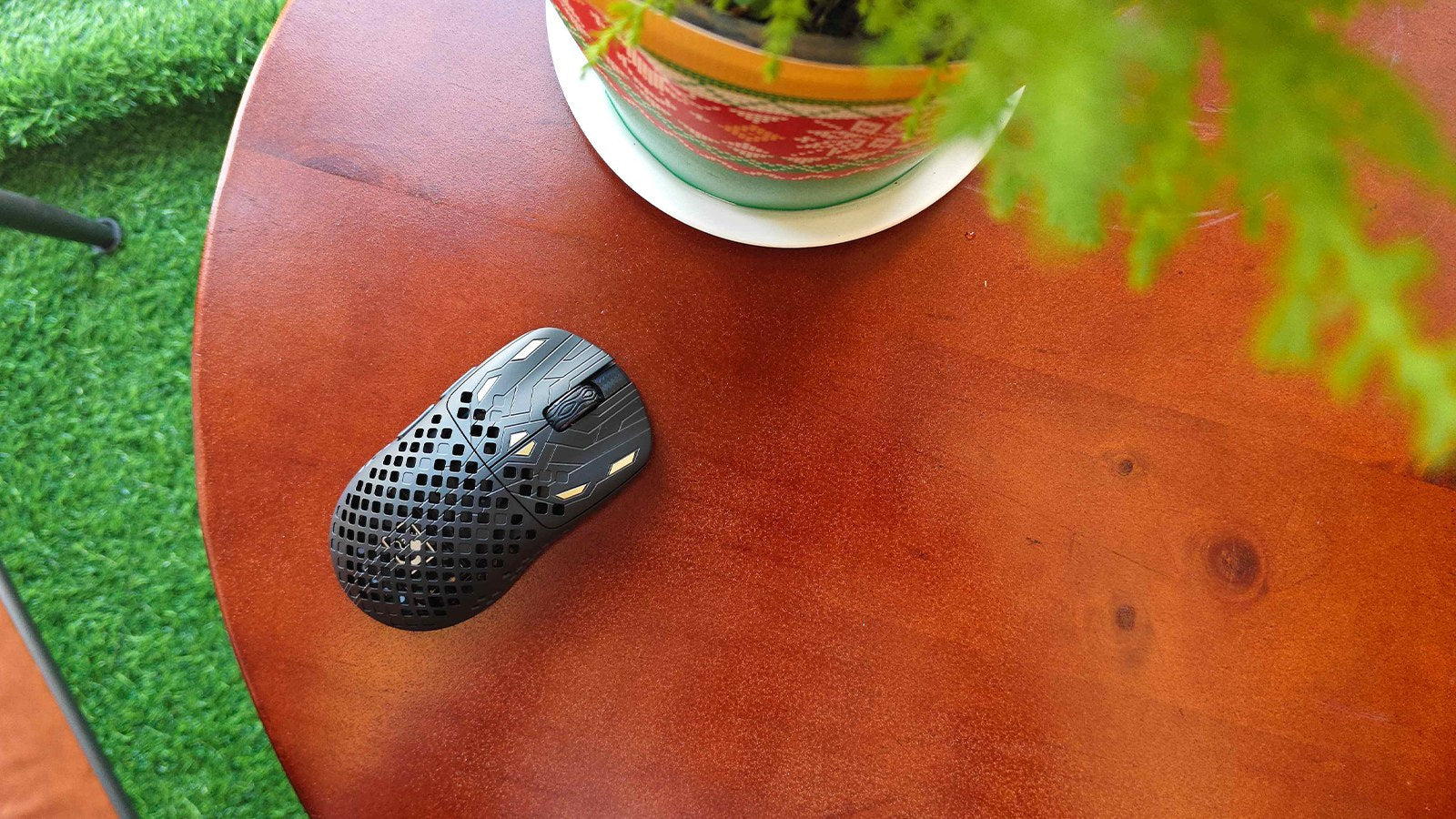 A black lightweight gaming mouse with a honeycomb shell, placed on a wooden table next to a potted plant