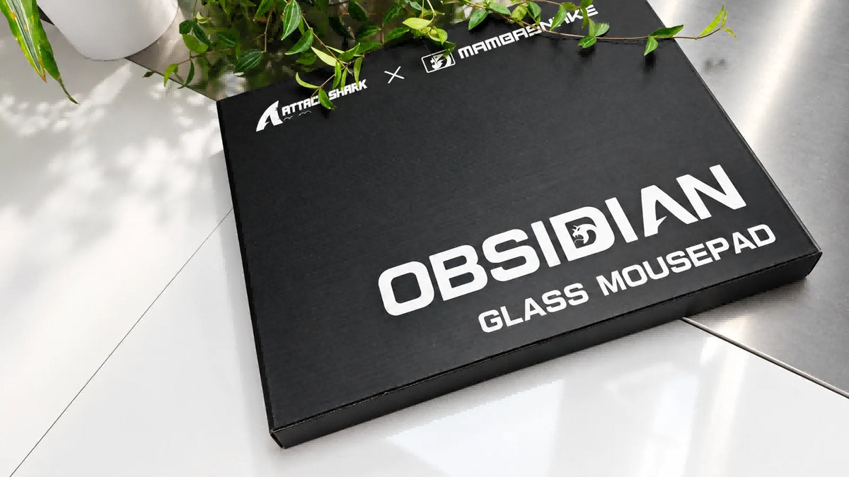 OBSIDIAN GLASS MOUSEPAD box on glossy white surface with green plant leaves.